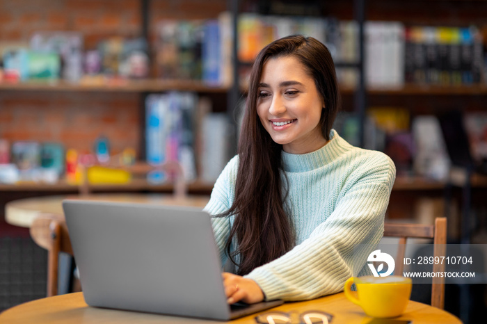 Pretty latin woman working on laptop computer in cafe, looking and smiling at screen, copy space