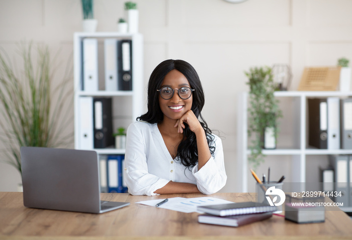 Positive African American businesswoman sitting at her desk with laptop computer in modern office