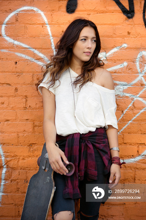 Young female posing with skateboard against brick wall with graffiti