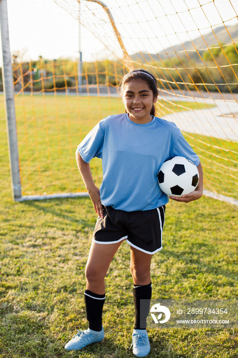 Portrait of soccer player with hand on hip holding ball while standing against net