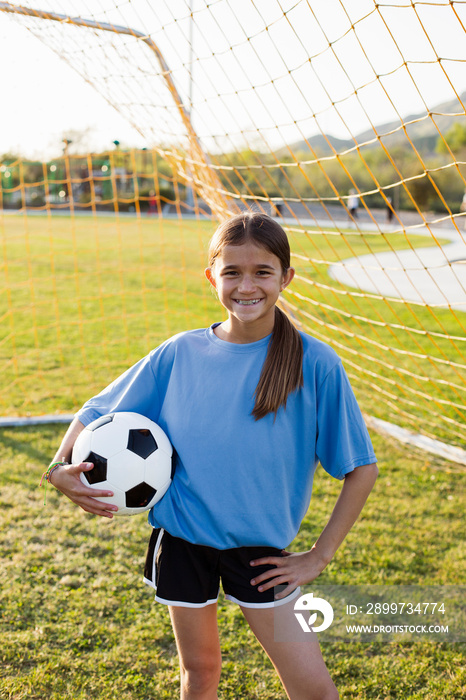 Portrait of cheerful girl holding soccer ball while standing on grassy field against net