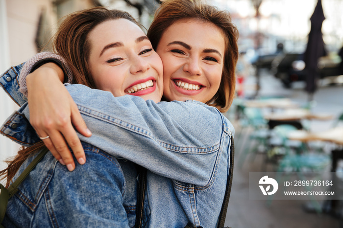 Close-up photo of two attractive happy woman friends hugging each other on city street, looking at c