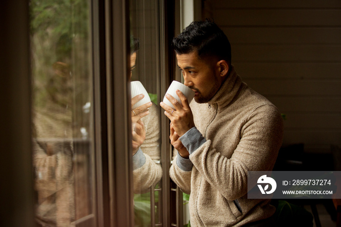 Thoughtful man drinking coffee looking through window while standing at home