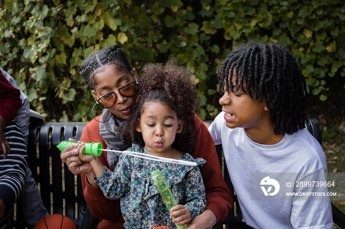 Young Black girl blowing bubbles with her family on bench