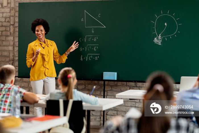 Happy African American math teacher explaining lecture on blackboard in the classroom.