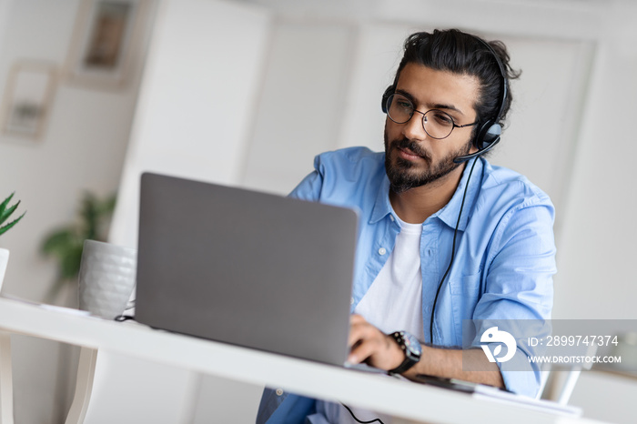 年轻的阿拉伯男性热线接线员戴着耳机在办公室里用笔记本电脑工作