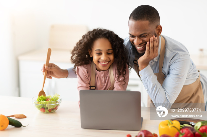 Black man and girl cooking in the kitchen using laptop