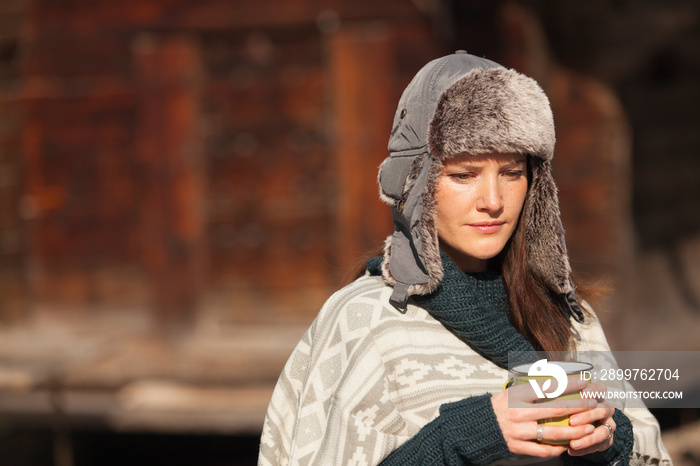 Thoughtful woman in fur hat drinking coffee outside cabin