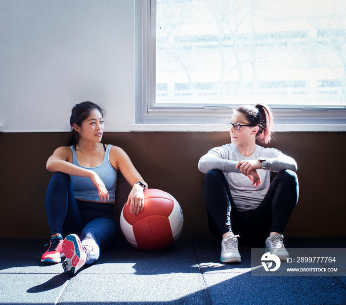 Full length of female athletes talking while sitting on floor against windows in gym