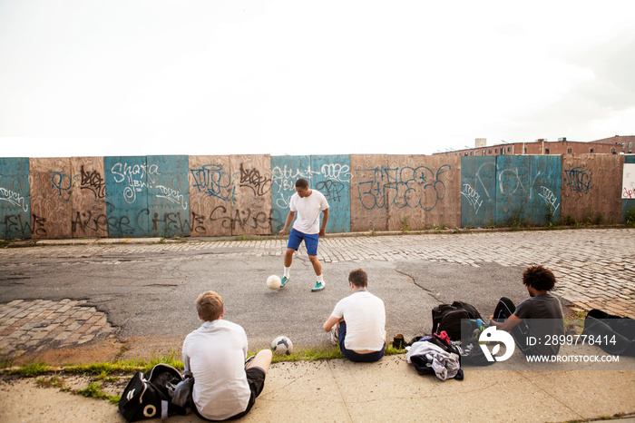 Man juggling ball while friends sitting on footpath against sky