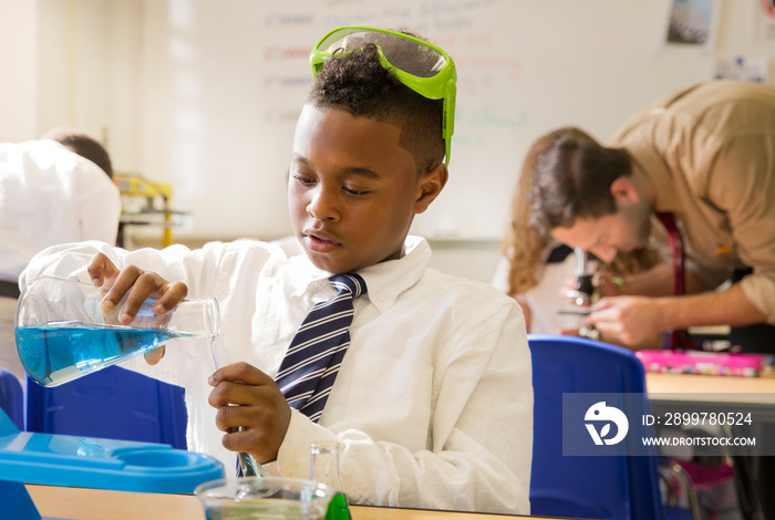 Portrait of schoolboy (8-9) sitting at desk in classroom pouring blue liquid from beaker to test tub