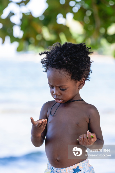 Retrato vertical de un pequeño niño afroamericano sin camisa de pie en la playa en un hermoso día so