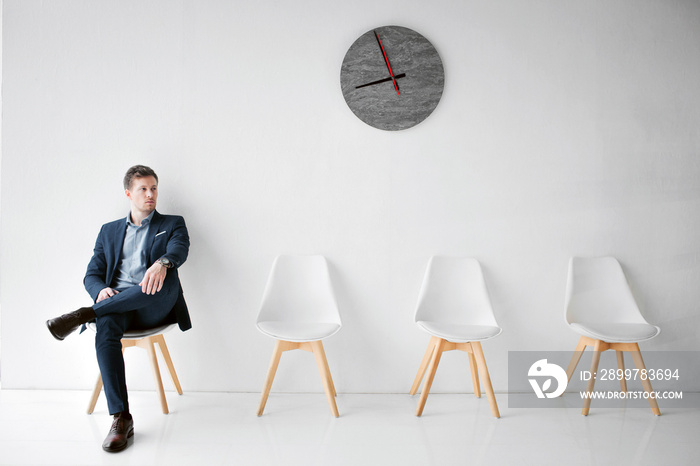 Serious young man in suit sit on white chair and look to right. He hold one leg on another. Guy wait