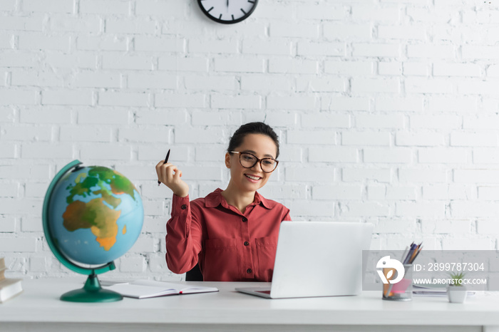 happy teacher in eyeglasses using laptop and holding pen while working from home.