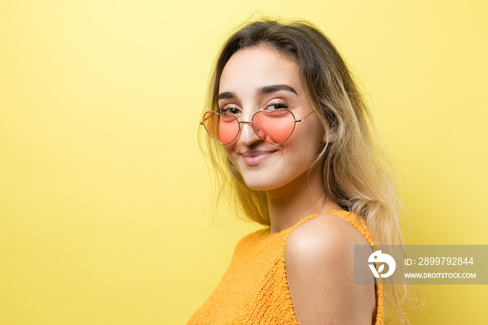 Romantic blonde-haired girl in trendy heart glasses posing with shy smile. Studio close-up portrait