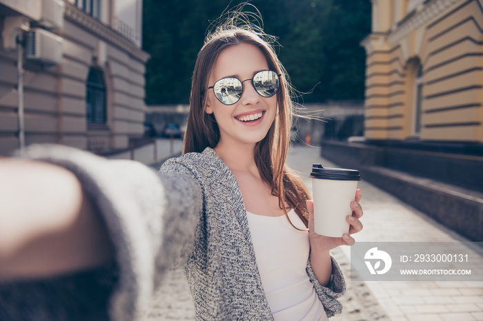 Cheert! Selfie time! Young happy lady in a spring vacation, walking in the city, drinking coffee, photographing herself on a sunny day