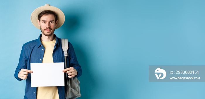 Image of smiling guy in straw hat backpacking, hitchhiking with piece of paper, travelling abroad on summer vacation, blue background