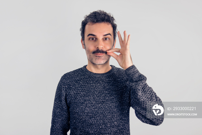 Funny portrait of brown, smiling, handsome man touching his mustache with gray sweater. Gray background.