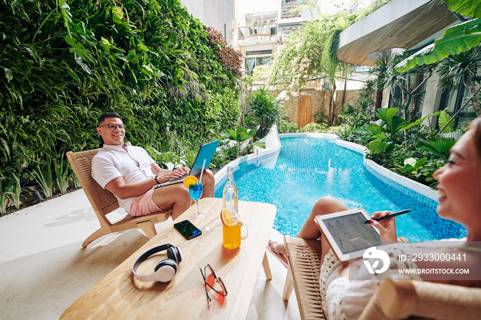 Happy man sitting in armschair by swimming pool, working on laptop and looking at his wife drawing fashion sketch on digital tablet