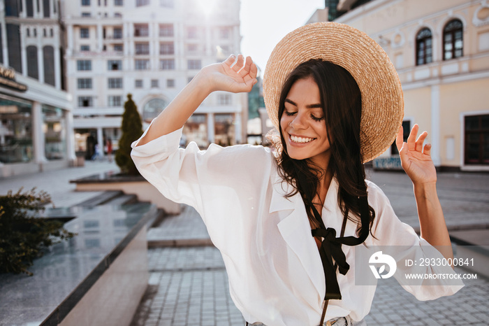 Good-humoured brunette lady posing on city background in hat. Outdoor portrait of attractive dark-haired girl standing on the street.