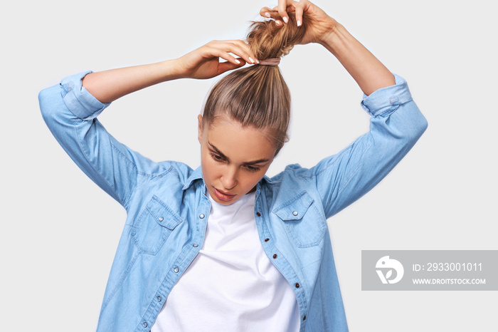 Indoor portrait of young woman collecting hair in a ponytail, wearing blue denim shirt and white t-shirt, posing over white wall. Adorable blonde female makes ponytail, advertises healthy natural hair