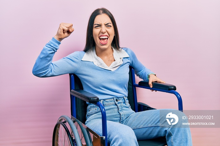 Beautiful woman with blue eyes sitting on wheelchair angry and mad raising fist frustrated and furious while shouting with anger. rage and aggressive concept.