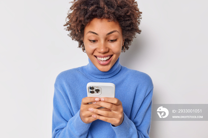 Happy teenage girl with dark curly hair uses shopping website on modern device sends sms smiles happily wears casual blue jumper isolated over white background. People and technologies concept