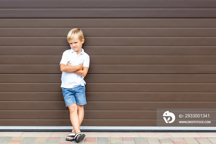 Cute grumpy blond child in casual clothing standing against brown garage door. Angry kid boy with crossed arms near house.Awkward age and parenting concept. Children and parents relations