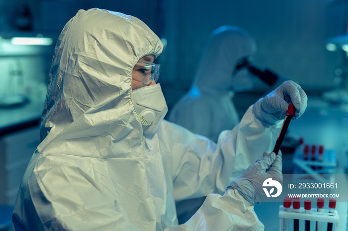 Scientist or chemist in coveralls holding vacutainer with biomaterial