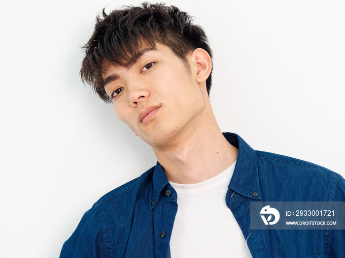 Portrait of handsome Chinese young man with curly black hair in blue shirt posing against white wall background. Looking at camera with tired expression, looks exhausted.