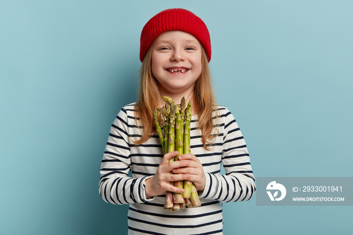 Organic vegetables concept. Cheerful positive lovely girl with green asparagus, smiles broadly, dressed in fashionable clothes, eats healthy food containing vitamins, isolated on blue background