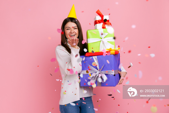 Extremely happy woman celebrating birthday holding stack of presents, standing under glitter rain, wearing white casual style sweater. Indoor studio shot isolated on pink background.