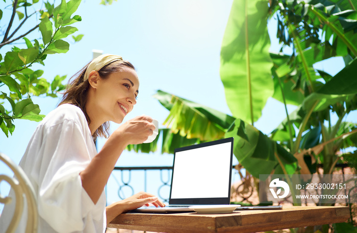 Cheerful freelancer womam drinking cofee at morning at balcony with palm trees, sitting with laptop blank screen. Work and vacation