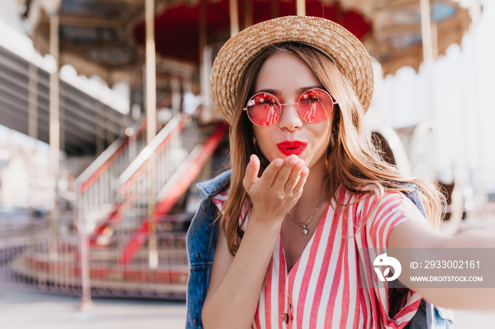 Pleasant caucasian woman sending air kiss while making selfie in straw hat. Outdoor photo of cheerful blonde girl in pink sunglasses taking picture of herself near carousel.