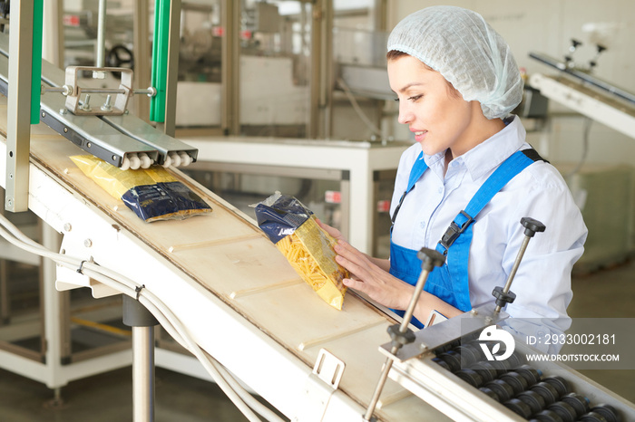 Portrait of female factory worker controlling  packaging process at modern food factory and looking at macaroni bags sliding down conveyor belt, copy space