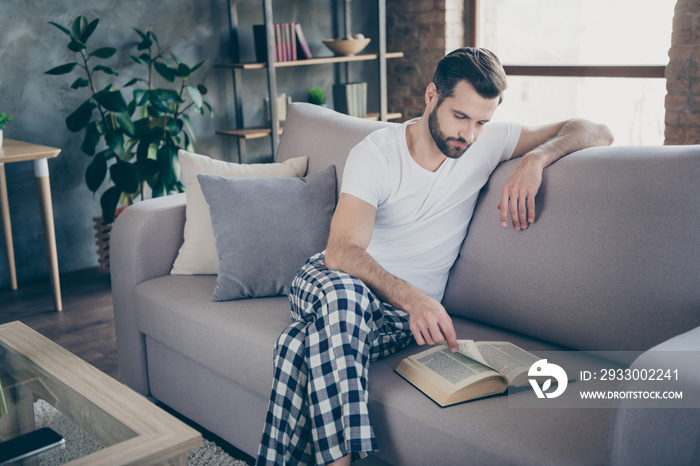 Portrait of his he nice attractive focused bearded guy sitting on divan reading interesting paper book page print spending free time leisure at modern loft industrial interior style indoors