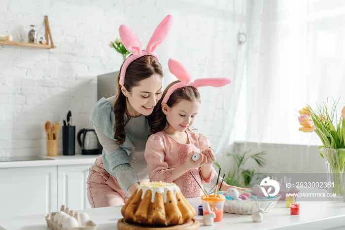 Happy mother and daughter in bunny ears looking at chicken eggs near tulips and easter bread