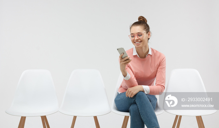 Portrait of european female student dressed casually holding mobile phone, typing messages, communicating with friends via social networks, using high-Internet connection sitting in the line