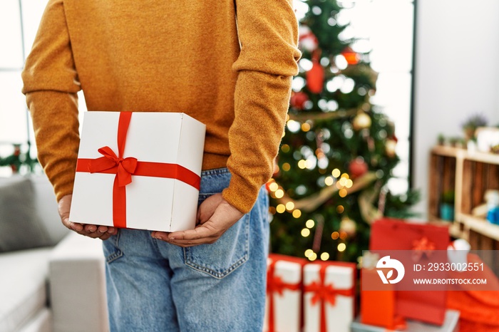 Man holding gift on his back standing by christmas tree at home.