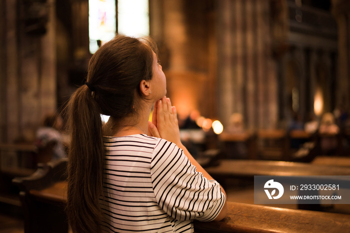 Religious young woman sitting on the bench in catholic church praying, looking up during church rule