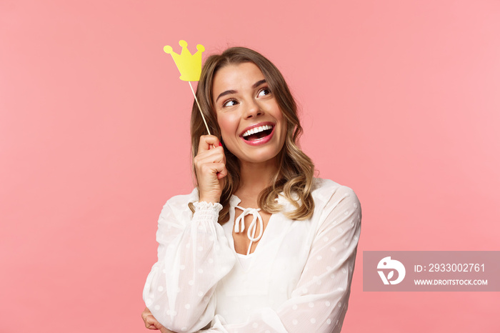 Spring, happiness and celebration concept. Close-up portrait of dreamy beautiful young blond girl imaging something cute and romantic, smiling look up daydreaming with crown on stick, pink background