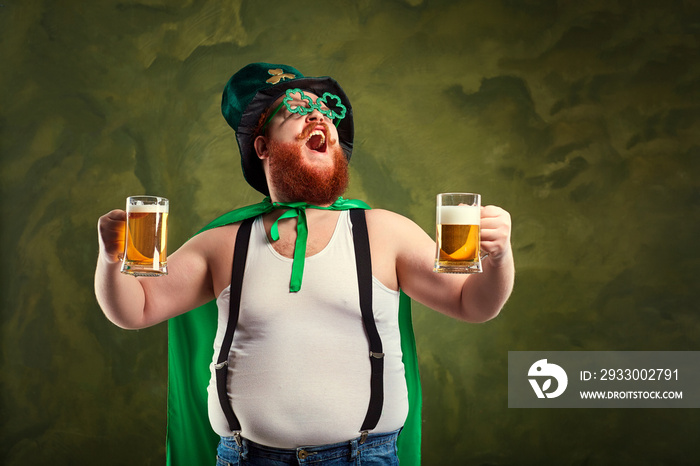 A fat man with a beard in St. Patrick’s suit is smiling with a mug of beer on a green background.