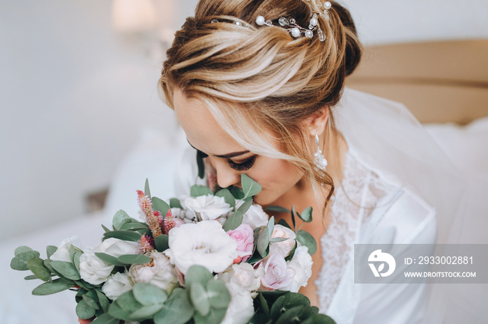 Blonde bride sits on a white bed and sniffs a bouquet. Wedding interior. Charges the bride at the hotel. Wedding photography. Portrait of a cute bride.