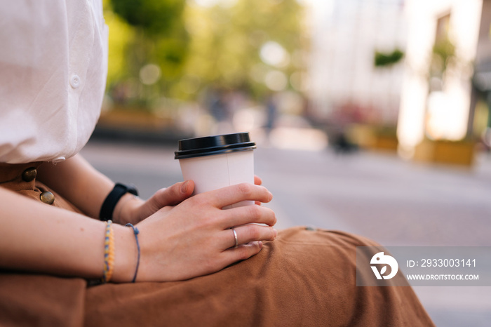 Close up side view cropped shot of unrecognizable elegant lady in casual clothes holding in hands paper cup with takeaway coffee sitting on bench on city street, in summer day, blurred background.