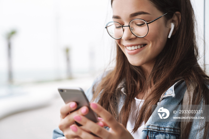 Smiling young pretty woman student wearing eyeglasses walking outdoors listening music with earphones using mobile phone chatting.