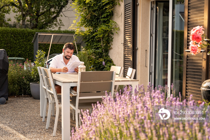 Adult man working or studying sitting on terrace in garden outdoors reading papers. Distant work, online studying. Slow living in village on nature.