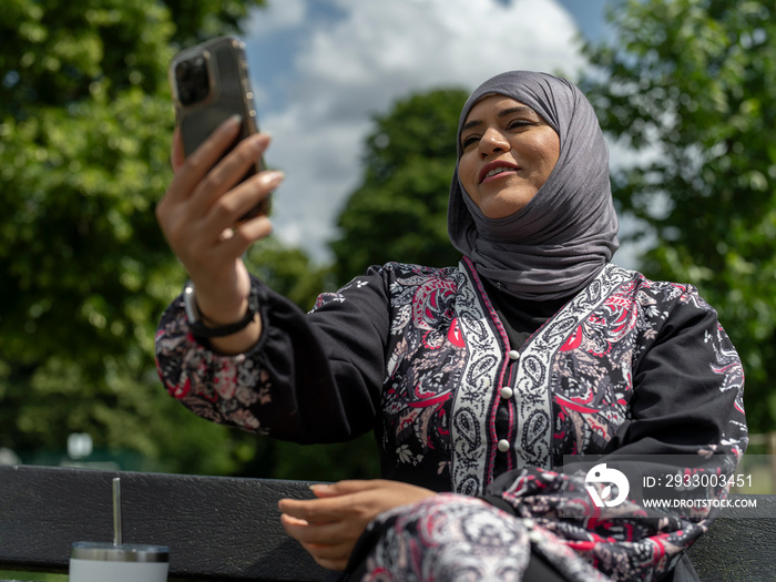 UK,Sutton,Woman in headscarf sitting on bench in park,talking on smart phone