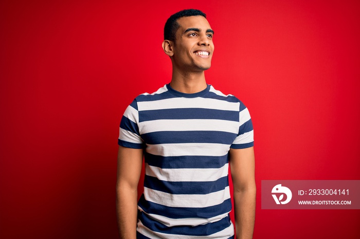 Handsome african american man wearing casual striped t-shirt standing over red background looking away to side with smile on face, natural expression. Laughing confident.