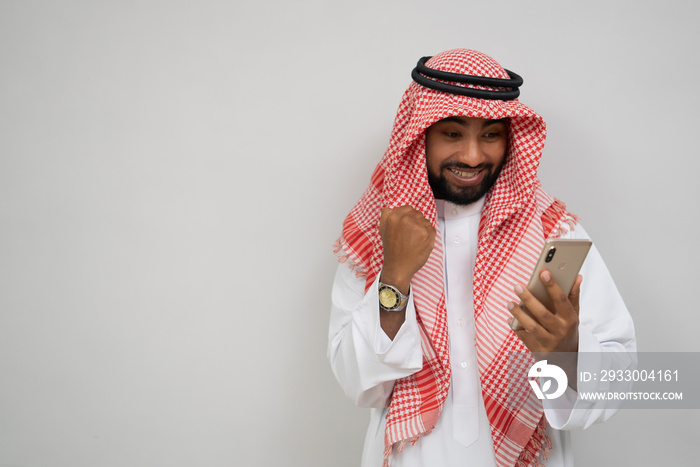 an arabian young man in a turban using a mobile phone happily looking at the screen with an excited expression on a plain background