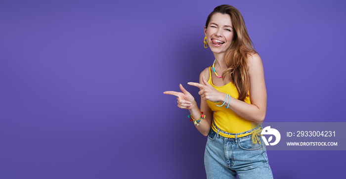 Playful young woman pointing away and making a face while standing against purple background